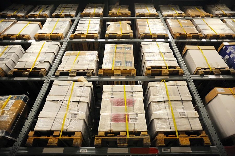 Stacks of shrink-wrapped pallets with boxes are stored on tall metal shelves in a warehouse, seen from a low angle. Woven polyester cord lashing, a flexible alternative for shock-sensitive loads, secures the pallets, and the shelves are organized and full.