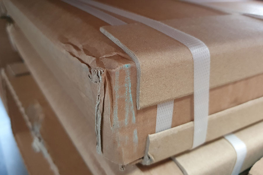 Close-up of a stack of cardboard boxes secured with white plastic straps. The corner of the top box shows strap damage, highlighting the risk of product loss during transit.
