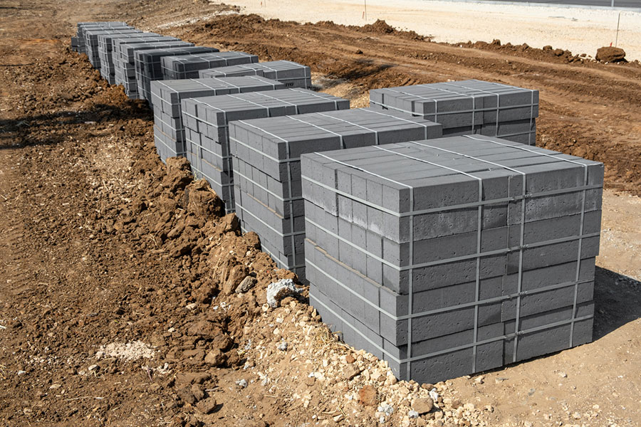 Stacks of gray concrete blocks are arranged in rows on a dirt construction site, secured with steel banding and ready for use as essential building materials.