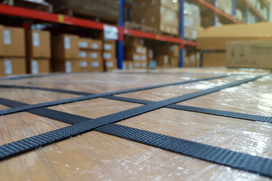 Close-up of a pallet wrapped in plastic and secured with durable PET strapping in a warehouse, with stacks of cardboard boxes and shelving visible in the background.