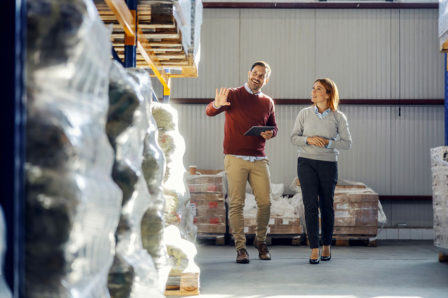 Two people walk in a warehouse, surrounded by pallet-wrapped goods secured with polypropylene strapping—an economical choice for light to medium loads. One person holds a tablet and gestures while speaking, and the other listens attentively. Both are dressed in business casual attire.